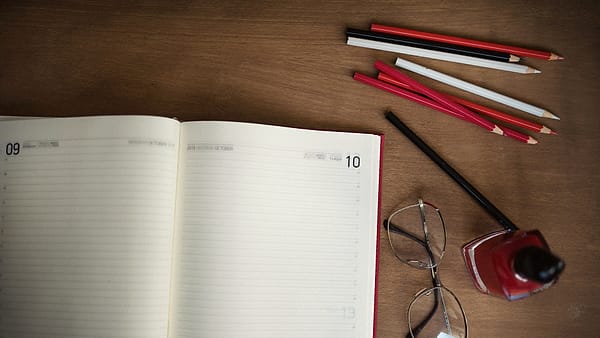 An open planner and colored pencils arranged on a wooden desk beside stationery