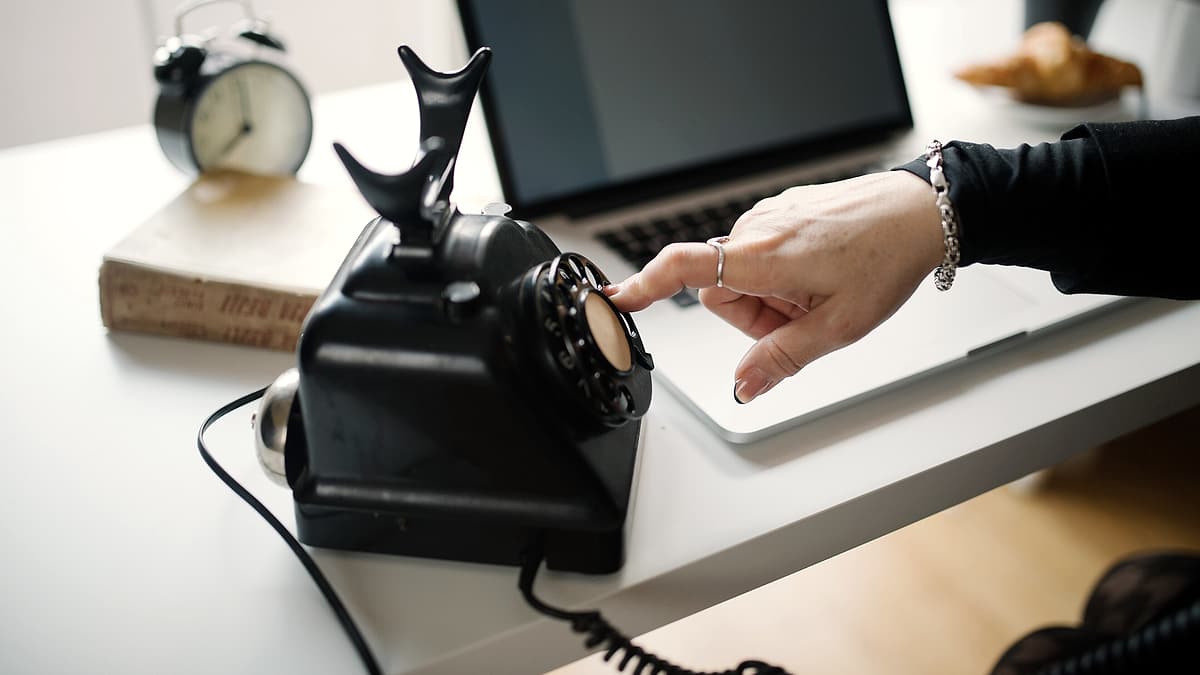 A hand dialing a rotary telephone standing on a desk beside a laptop